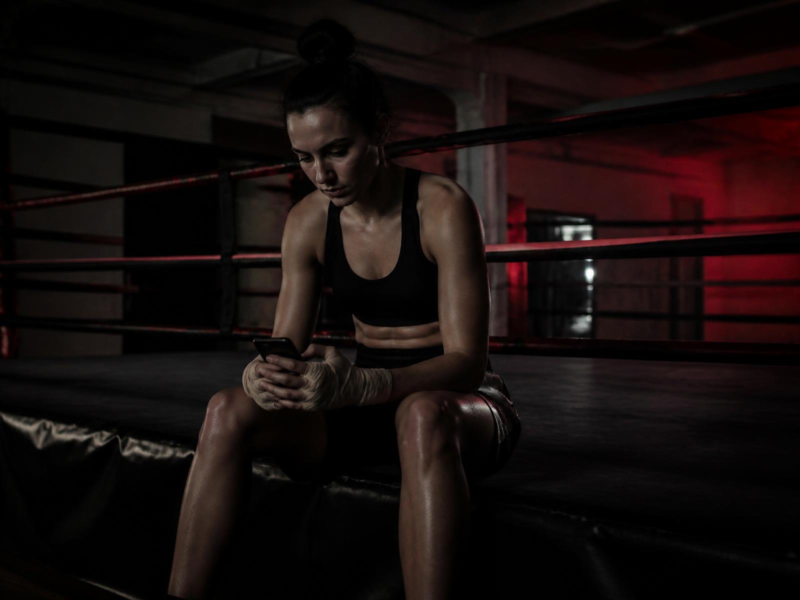 Fighter sitting on the ring apron of an empty gym at night looking at a phone